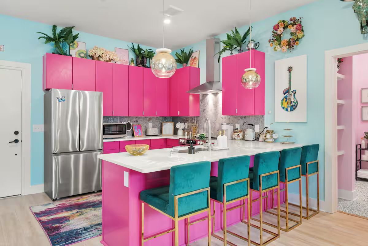 Colorful modern kitchen interior with hot-pink cabinets, marble island and breakfast bar, teal velvet bar stools with gold frames, stainless fridge, pendant lights, plants and wall art.