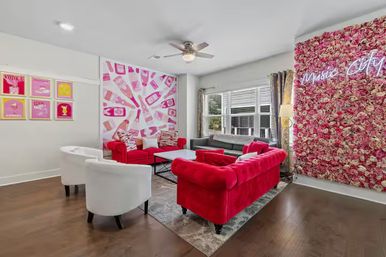 Urban living room lounge with red velvet tufted sofas, white club chairs, playful pink pop-art wall and floral neon 'Music City' accent wall, large window and hardwood floor.