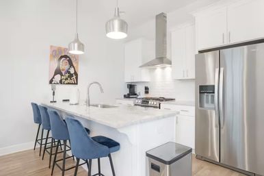 Bright modern white kitchen with marble island and undermount sink, row of blue velvet bar stools, stainless steel refrigerator and range with hood, pendant lights and colorful wall art.