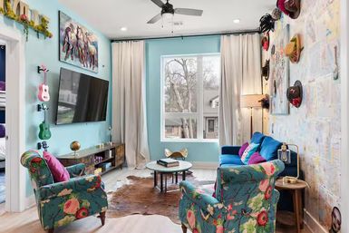 Cheerful turquoise living room in a suburban home with floral armchairs, a blue sofa, wall-mounted TV, hat gallery wall, small guitars, cowhide rug, and a large window letting in daylight.