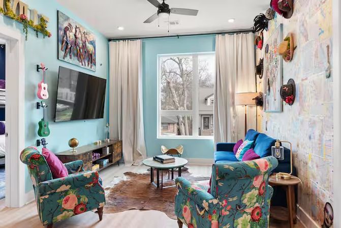 Cheerful turquoise living room in a suburban home with floral armchairs, a blue sofa, wall-mounted TV, hat gallery wall, small guitars, cowhide rug, and a large window letting in daylight.
