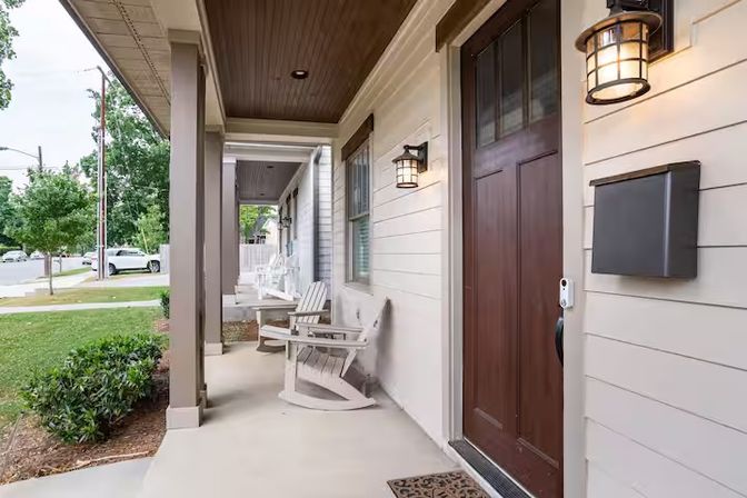 Inviting suburban front porch with dark wood door, white Adirondack rocking chairs, lantern wall lights, mailbox and concrete stoop overlooking a tree-lined residential street