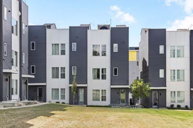 Sleek gray-and-white three-story townhomes facing a shared grassy courtyard with young trees and front entrances under a blue sky.