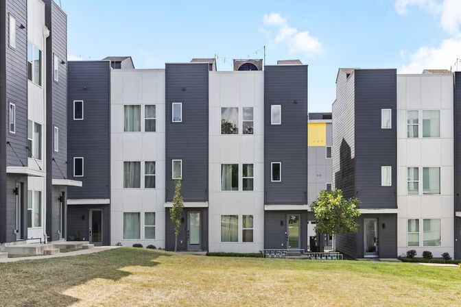 Sleek gray-and-white three-story townhomes facing a shared grassy courtyard with young trees and front entrances under a blue sky.