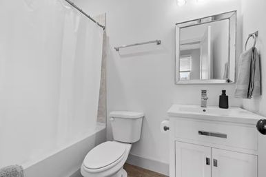 Clean compact residential bathroom with shower-tub and white curtain, toilet, modern white vanity and mirror, towel on ring and wood-look flooring.