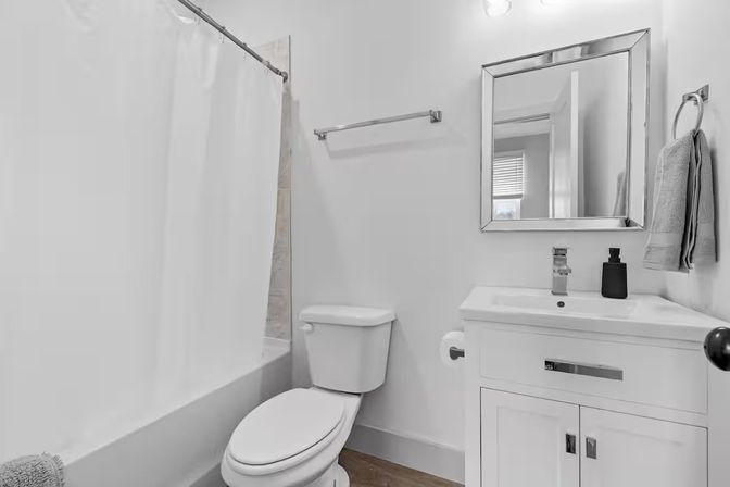 Clean compact residential bathroom with shower-tub and white curtain, toilet, modern white vanity and mirror, towel on ring and wood-look flooring.