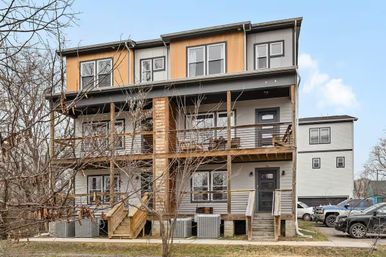 Three-story modern triplex with gray siding and orange upper panels, wooden balconies and stairs, leafless trees in front and cars parked in the driveway under a clear blue sky.