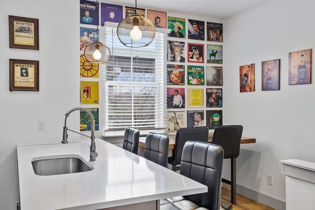 Modern kitchen dining nook with white quartz island and stainless sink, tall black leather bar stools, wire globe pendant lights, and a colorful retro vinyl album wall beside a window with white blinds.