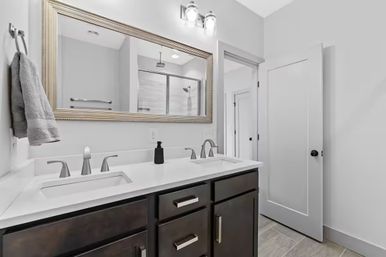 Bright modern residential bathroom with dark wood double vanity, white quartz countertop, twin chrome faucets, large framed mirror and glass-enclosed shower