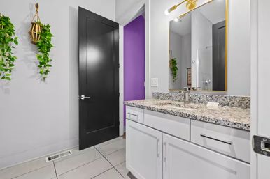 Contemporary bathroom interior with white cabinets and speckled granite vanity, gold-framed mirror and brass sconce, black door opening to a purple accent wall, hanging green plants and light gray tile floor.