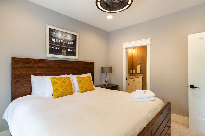 Cozy modern bedroom with wooden headboard, king bed in white linens and yellow accent pillows, bedside lamp, folded towels and doorway to en-suite bathroom.