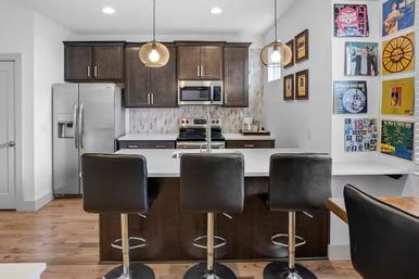 Sleek modern kitchen with white island and three black swivel bar stools, stainless steel fridge and oven, dark wood cabinets, pendant lights, hardwood floors, and colorful vinyl record wall art.