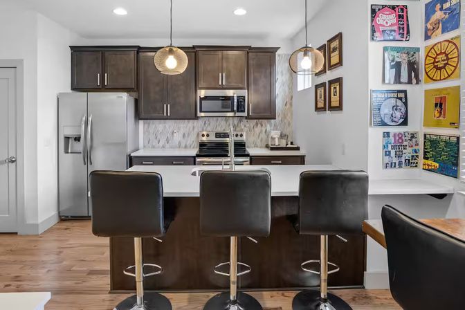 Sleek modern kitchen with white island and three black swivel bar stools, stainless steel fridge and oven, dark wood cabinets, pendant lights, hardwood floors, and colorful vinyl record wall art.