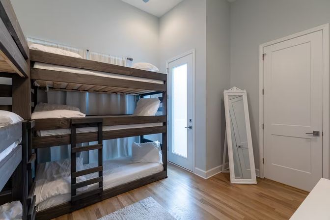 Bright modern bedroom with dark wooden stacked bunk beds dressed in crisp white linens, hardwood floors, a leaning full-length mirror, and a frosted-glass exterior door.