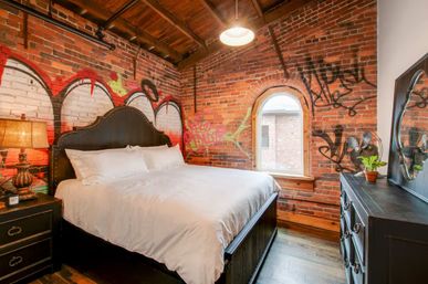 Edgy urban loft bedroom with exposed graffiti-covered brick walls, king bed, arched window, industrial pendant light and dark wood furniture.