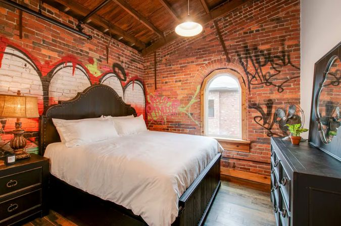 Edgy urban loft bedroom with exposed graffiti-covered brick walls, king bed, arched window, industrial pendant light and dark wood furniture.