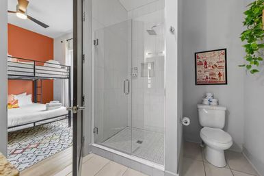 Compact modern bathroom with clear glass shower, white toilet topped with stacked toiletries and a hanging plant; open door reveals adjacent bedroom with black metal bunk bed, orange accent wall and patterned rug in a stylish urban rental.