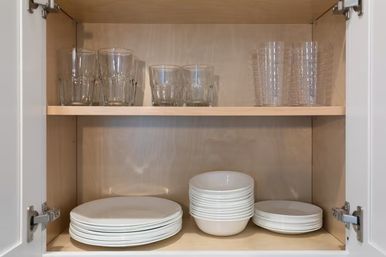 Neat kitchen cabinet interior showing light wood shelves with stacked white plates and bowls below and clear drinking glasses on the upper shelf