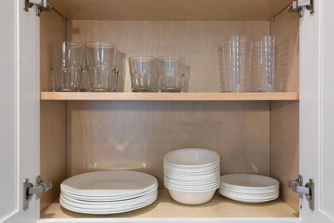 Neat kitchen cabinet interior showing light wood shelves with stacked white plates and bowls below and clear drinking glasses on the upper shelf