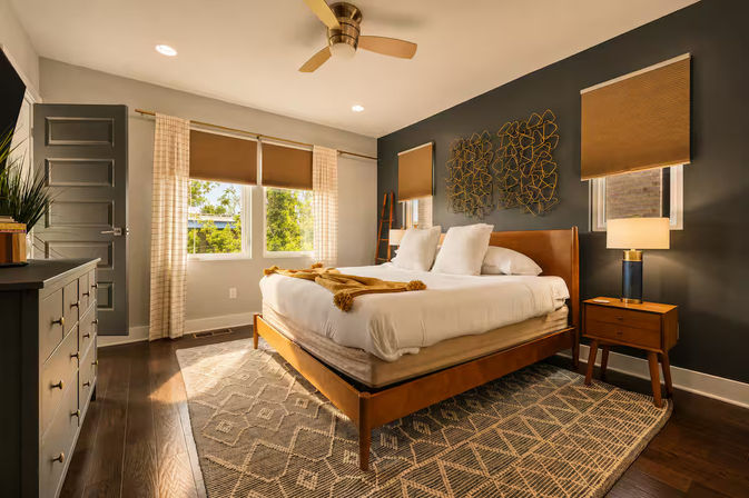 Sunlit modern bedroom with a mid-century wooden bed dressed in white linens and mustard throw, dark gray accent wall with metal art, bedside lamp on a wooden nightstand, patterned area rug, hardwood floors, ceiling fan, and leafy window view.