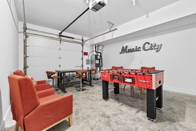 Finished garage turned game room: red foosball table, cozy orange lounge chairs, bar-height table with stools, concrete floor and a 'Music City' wall sign.
