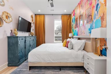 Stylish modern apartment bedroom with bold abstract mural accent wall, mustard-gold curtains framing a window, teal blue dresser with wall-mounted TV, white bed and nightstand, and colorful throw pillows.