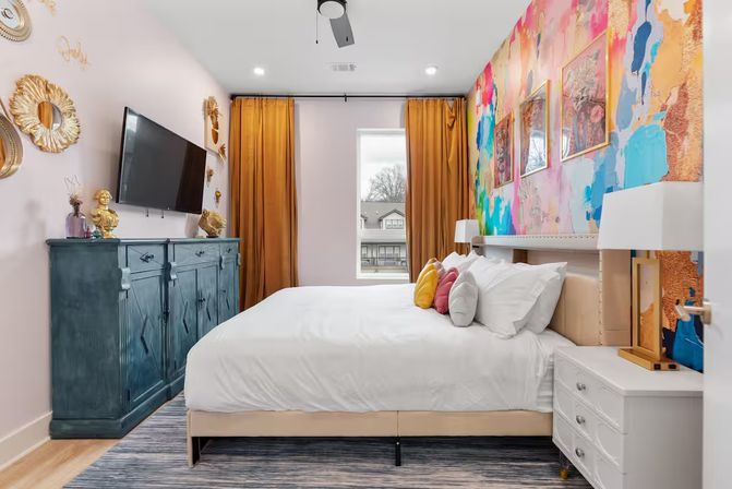 Stylish modern apartment bedroom with bold abstract mural accent wall, mustard-gold curtains framing a window, teal blue dresser with wall-mounted TV, white bed and nightstand, and colorful throw pillows.