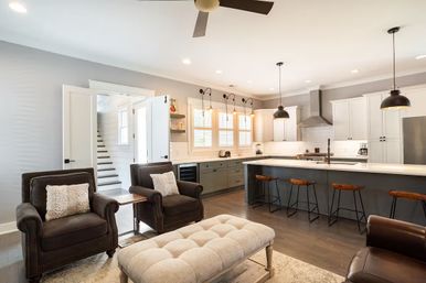 Bright modern farmhouse open-concept kitchen and living room with a gray island, white cabinets, industrial pendant lights, wooden bar stools, leather armchairs and a tufted ottoman on hardwood floors.