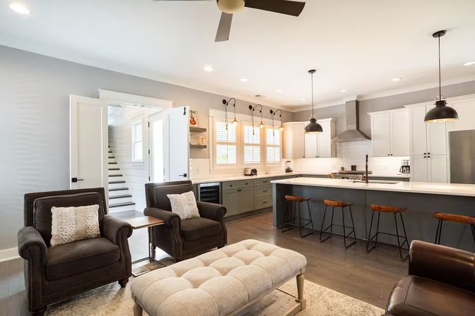 Bright modern farmhouse open-concept kitchen and living room with a gray island, white cabinets, industrial pendant lights, wooden bar stools, leather armchairs and a tufted ottoman on hardwood floors.