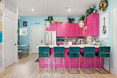 Vibrant modern open-plan kitchen with hot-pink cabinets, white quartz island, five teal velvet bar stools with gold frames, stainless steel appliances, globe pendant lights, aqua walls, potted plants and colorful wall art.