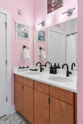 Pastel pink bathroom with a large mirror above a white double-sink countertop, matte black faucets, wood vanity cabinets, soap dispensers and framed floral art.
