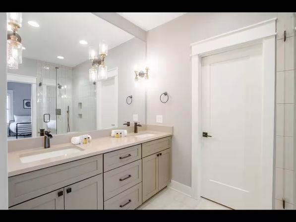 Bright contemporary master bathroom with double sink vanity, gray shaker cabinets, beige quartz countertop, large wall mirror, brass glass sconces, and glass-enclosed shower reflected in mirror.