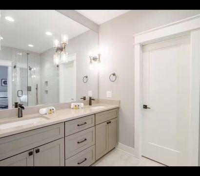 Bright contemporary master bathroom with double sink vanity, gray shaker cabinets, beige quartz countertop, large wall mirror, brass glass sconces, and glass-enclosed shower reflected in mirror.