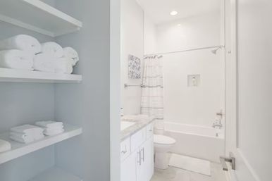 Bright modern residential bathroom with white bathtub and striped shower curtain, marble-top vanity, and open shelves stacked with rolled white towels for a clean spa-style look.