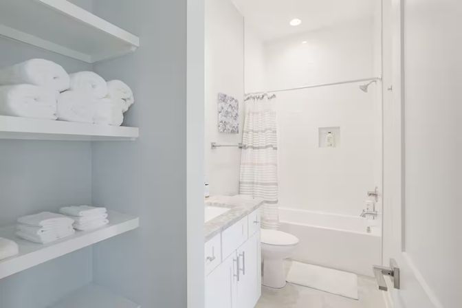 Bright modern residential bathroom with white bathtub and striped shower curtain, marble-top vanity, and open shelves stacked with rolled white towels for a clean spa-style look.