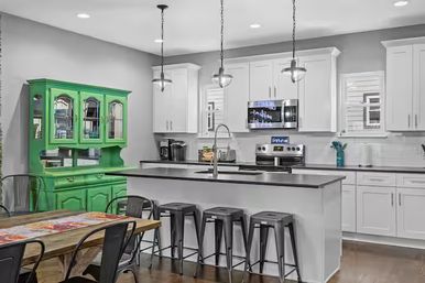 Open-concept modern kitchen with white cabinets and stainless steel appliances, large island with black countertop and five metal stools, pendant lights, wooden dining table and bright green vintage hutch.