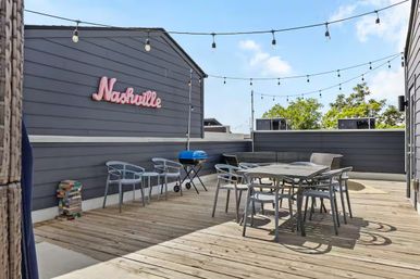 Sunny Nashville rooftop deck with wood flooring, hanging string lights, pink Nashville sign, outdoor dining table and chairs, and a small blue grill.