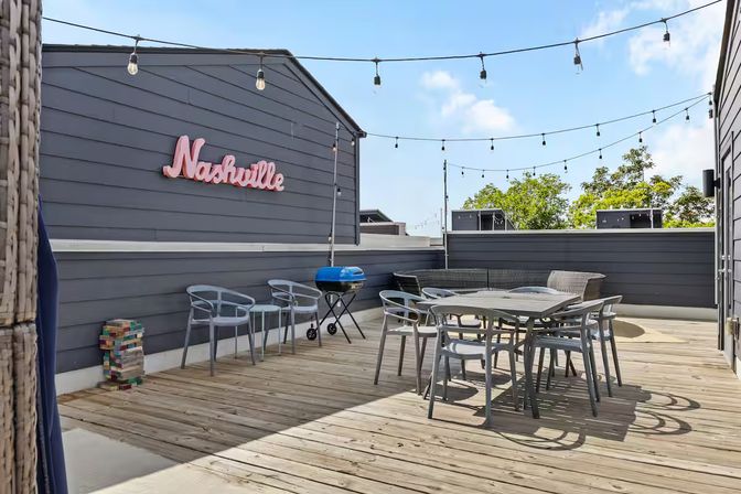 Sunny Nashville rooftop deck with wood flooring, hanging string lights, pink Nashville sign, outdoor dining table and chairs, and a small blue grill.