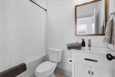Compact modern residential bathroom with white shower-tub and curtain, toilet, white vanity topped with folded gray towels and black soap dispenser, and a wood-framed mirror reflecting a window.