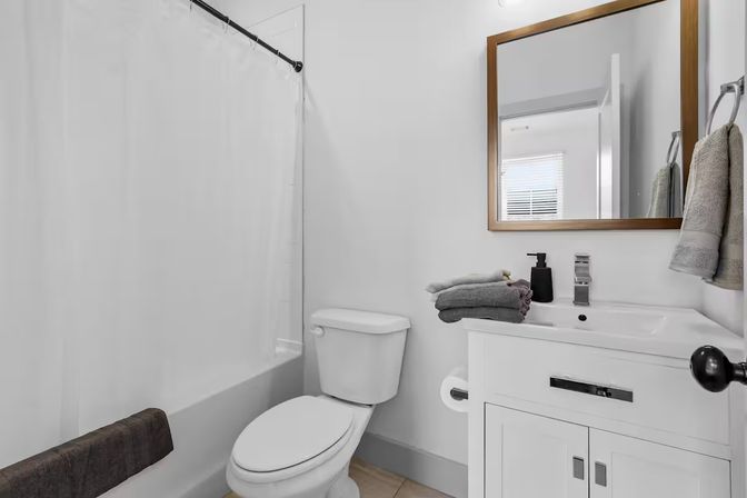 Compact modern residential bathroom with white shower-tub and curtain, toilet, white vanity topped with folded gray towels and black soap dispenser, and a wood-framed mirror reflecting a window.