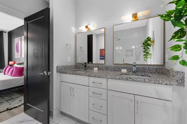 Contemporary apartment bathroom with double-sink granite vanity, white cabinets, gold-framed mirrors, hanging plant, and an open door showing a bedroom with bright pink pillows.