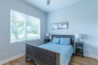 Bright airy bedroom with blue bedding on a dark wood bed, matching nightstands, white blinds on a large window and hardwood floors