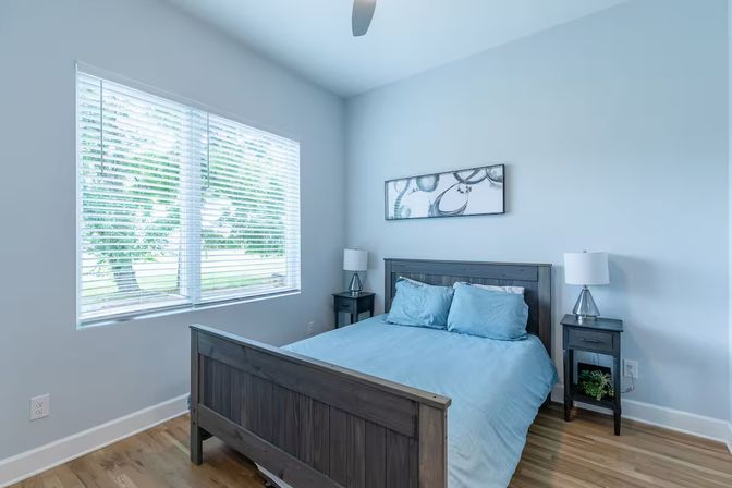 Bright airy bedroom with blue bedding on a dark wood bed, matching nightstands, white blinds on a large window and hardwood floors