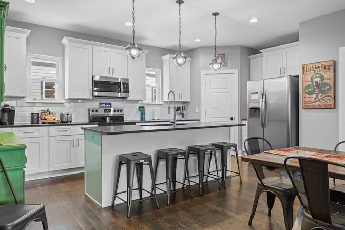 Bright open-concept suburban kitchen with white shaker cabinets, large island with black countertop and four metal bar stools, stainless steel refrigerator and range, globe pendant lights and dark hardwood floors.