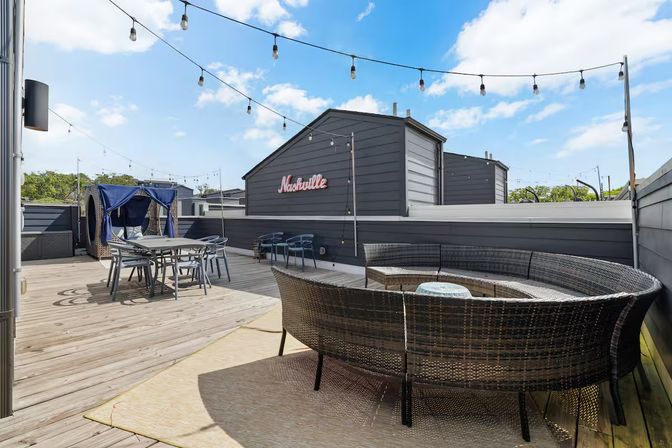Rooftop patio in Nashville with curved wicker lounge, outdoor dining table, blue-cushioned cabana, hanging string lights and gray siding under a bright blue sky