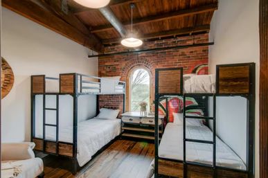 Industrial loft bedroom with two metal-framed bunk beds, exposed brick wall, arched window, wooden ceiling and hardwood floors