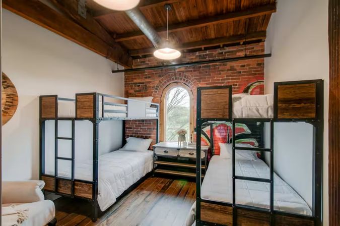Industrial loft bedroom with two metal-framed bunk beds, exposed brick wall, arched window, wooden ceiling and hardwood floors