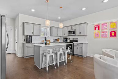 Modern open-plan kitchen with gray cabinets, white quartz island and three white metal bar stools, stainless steel appliances, crystal pendant lights, hardwood floors and colorful cocktail art on the wall.