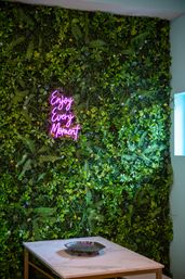 Indoor living green wall with ferns and small yellow flowers, cheerful pink neon sign reading "Enjoy Every Moment", marble-top table with decorative metal tray — modern interior plant wall decor.