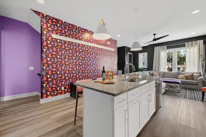 Contemporary open-concept kitchen and living room with playful cactus-pattern accent wall and purple entry wall, granite island with white cabinets and sink, pendant lights, hardwood floors, gray sectional sofa and large windows with curtains.